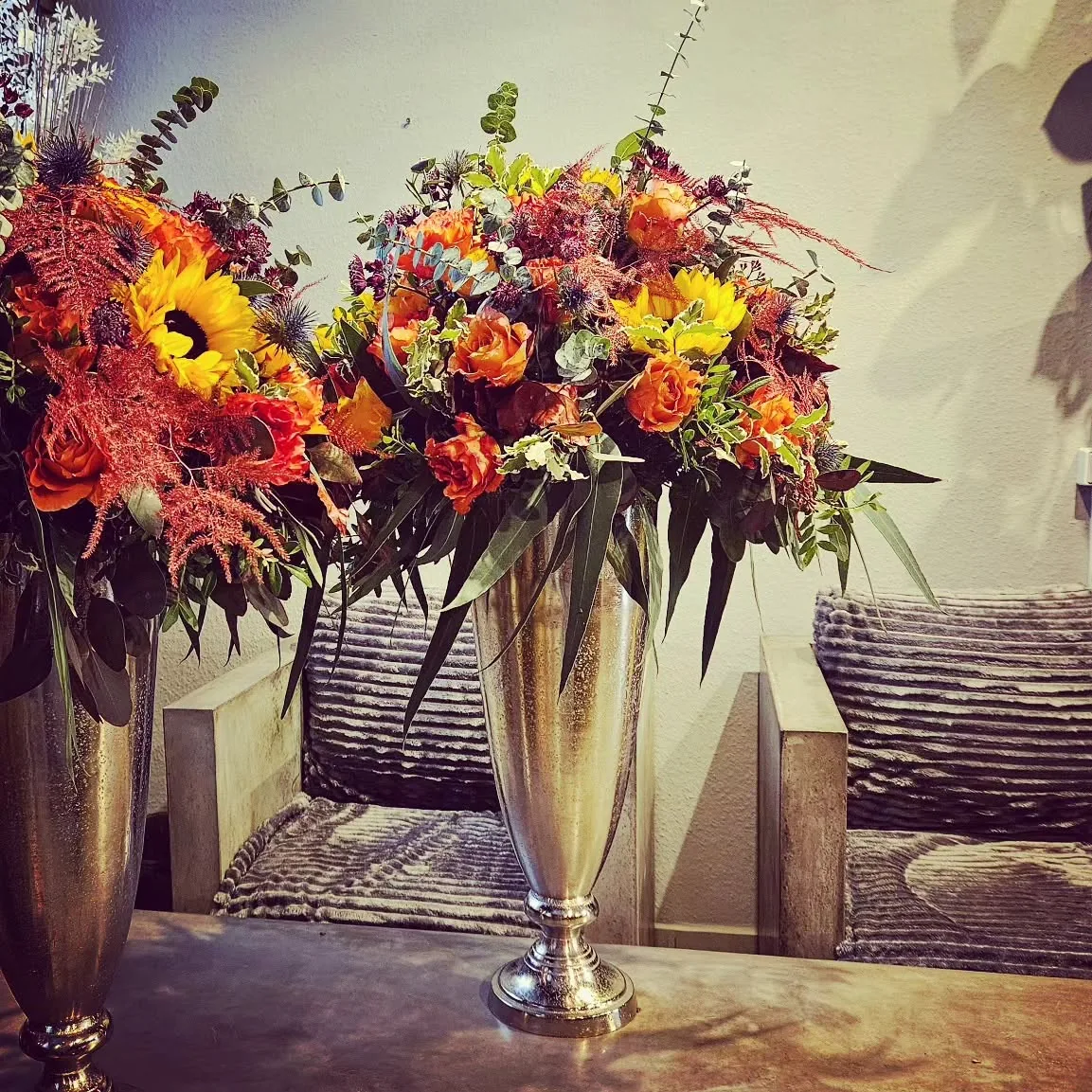 Farbenfrohe Eventfloristik mit orangefarbenen Rosen, Sonnenblumen und dunklen Akzenten in hoher silberner Vase